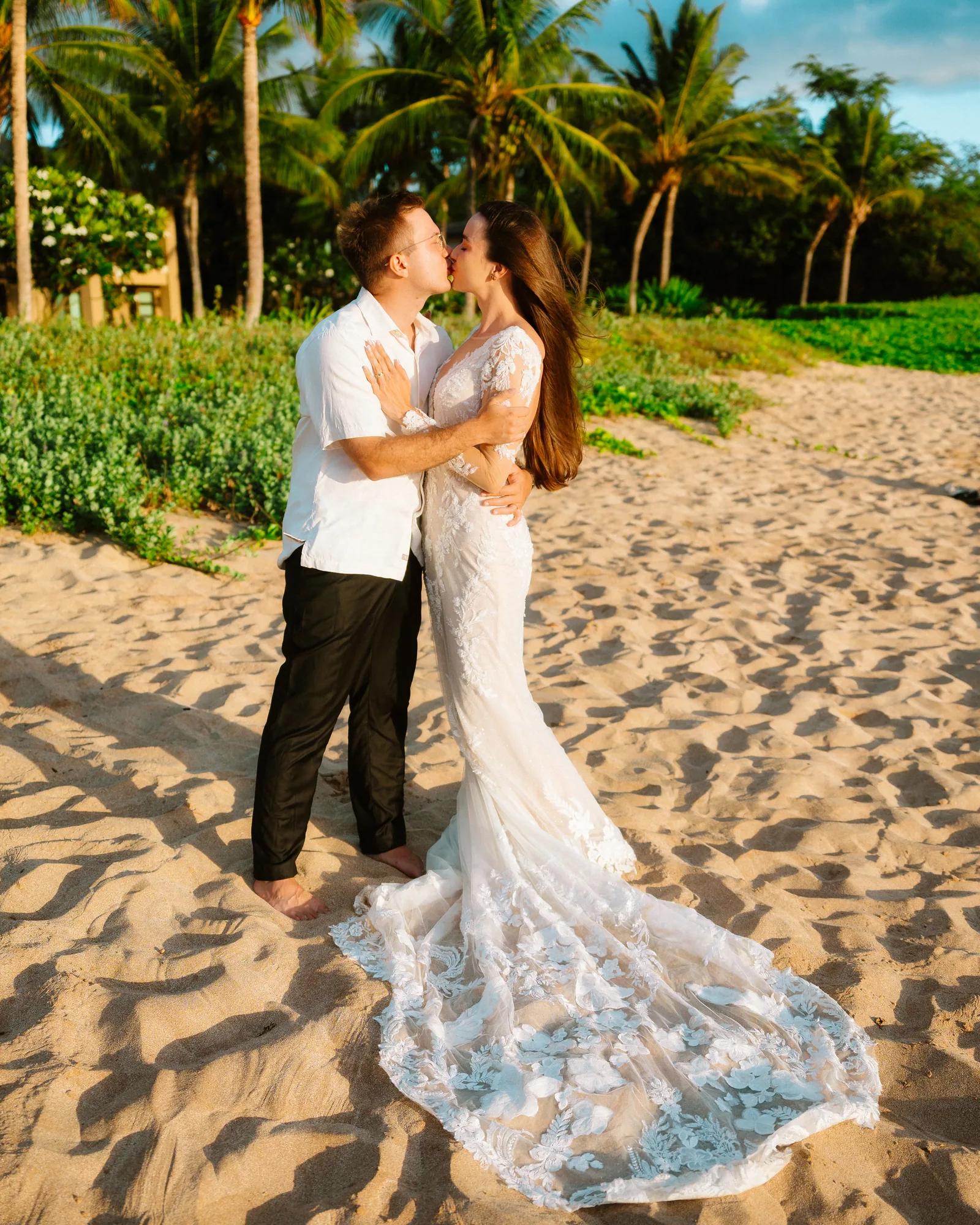 A bride in a lace gown with her partner on the sand beneath palms